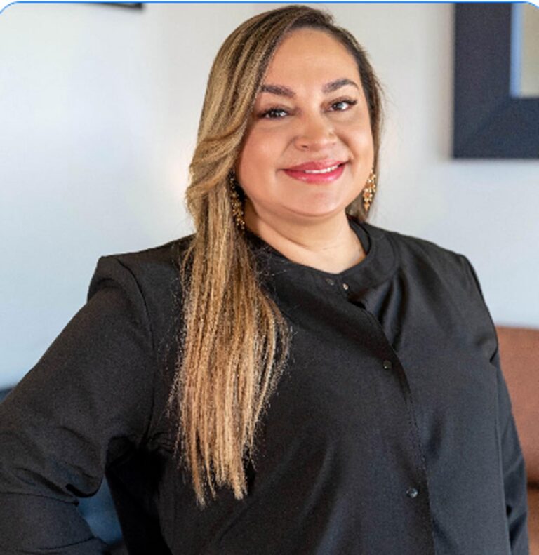 Smiling individual with long light brown hair and blonde highlights, wearing a black button-up shirt and gold earrings. Indoor setting includes a white wall with framed decor and part of a brown couch.