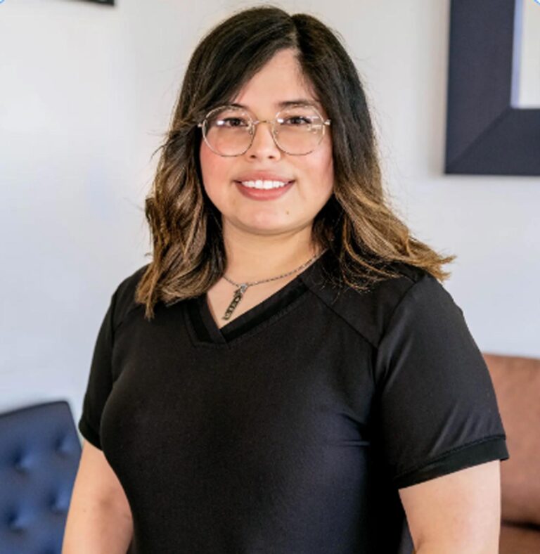 Smiling individual with shoulder-length wavy hair and lighter highlights, wearing glasses and a black V-neck shirt. A pendant necklace is visible, with a tufted chair and partial picture frame in the background.
