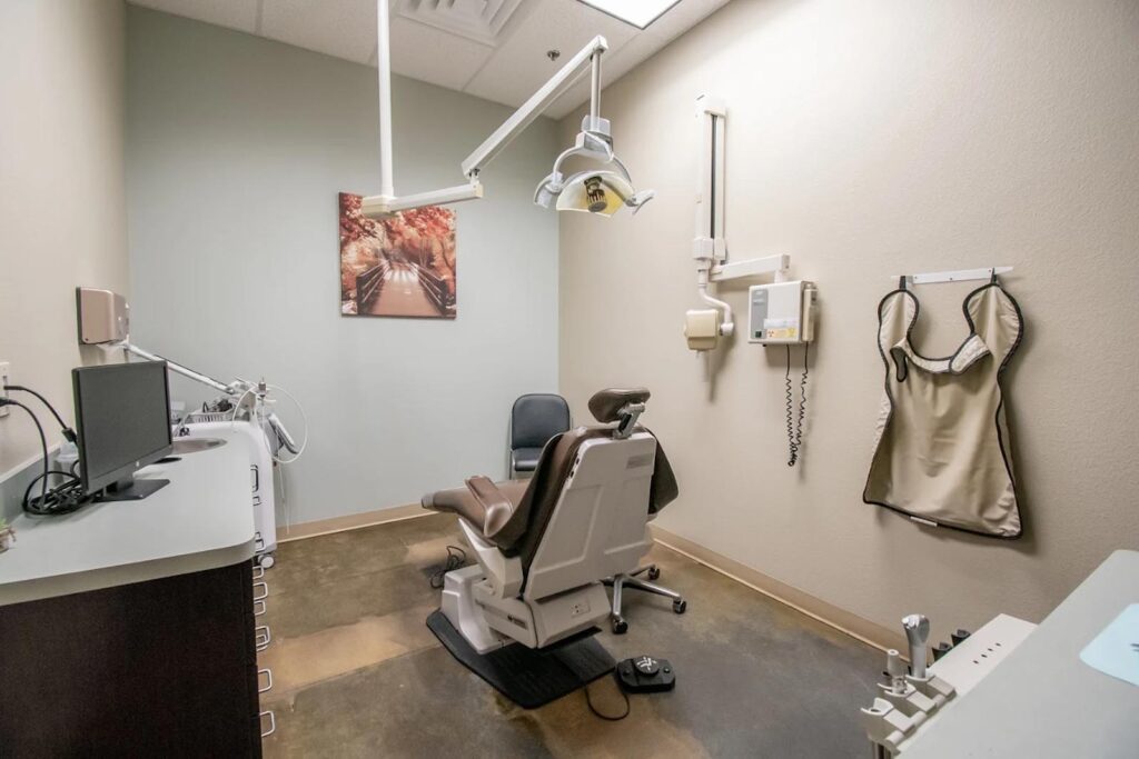 Dental exam room with a central chair, overhead light, and mounted equipment including an X-ray unit and lead apron. Clean layout includes a counter with instruments, a black guest chair, and a framed picture of a tree-lined path.